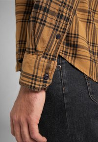 Camicia a quadri marrone e nera con orlo arrotondato e polsini abbottonati, realizzata in tessuto testurizzato, abbinata a jeans scuri.
