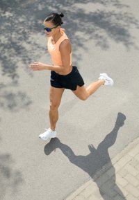 Běžecké boty, bílé s akcenty; oranžový bezrukávový top; černé šortky s vertikálními pruhy; stín vržený na chodník.