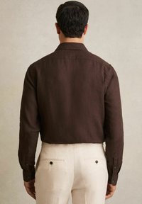 Camisa de lino marrón con mangas largas y puños abotonados, que presenta un cuello y un corte recto. Llevada con pantalones sastre de color claro.
