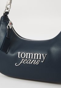 Navy blue handbag made of smooth material features a zip closure and a silver logo "tommy jeans" prominently displayed. Accented with silver hardware.