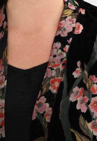 Black velvet garment with floral patterns in pink, red, and green. Features a shawl collar and a soft texture, partially shown over a black top.