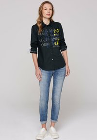 Maglietta nera a bottoni con motivi di testo colorati, abbinata a jeans blu chiaro e sneakers bianche, presentata su un modello.