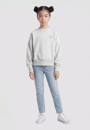 Fille avec une coiffure à double chignon portant un sweat-shirt gris clair, un jean bleu clair et des baskets montantes blanches, debout devant un fond blanc.