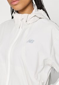 Casaco leve branco com colarinho alto, fecho éclair à frente e discreto logótipo da New Balance. Textura suave e design minimalista.