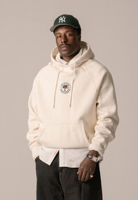 Cream-colored hoodie with a kangaroo pocket, featuring a circular tree logo. Worn over a white collared shirt, paired with black jeans.
