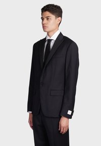 Giacca da smoking nera con risvolti in satin, chiusura a bottone singolo, due tasche frontali e camicia bianca sotto. Vestibilità su misura.