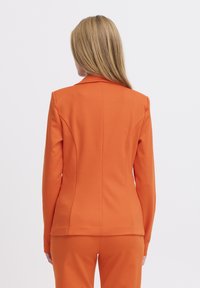Blazer fitted arancione con spalle strutturate e una texture liscia, caratterizzato da dettagli di cucitura lungo la schiena e colletto a rever.