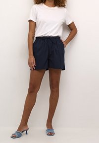 Personne portant une chemise blanche à manches courtes, un short bleu marine avec cordon de serrage, et des talons ouverts bleu clair effilochés, debout devant un fond clair uni.