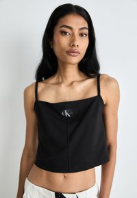 Svart cropped topp med tynne stropper, med firkantet utringning og et logo-patch foran, laget av et glatt materiale.