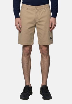 Homme portant un short beige jusqu'aux genoux avec un logo noir sur la jambe gauche, des chaussures noires et un haut à manches longues foncé, debout face à l'avant.