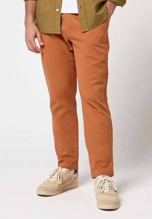 Homme portant un pantalon couleur rouille, des baskets beiges, des chaussettes blanches, une chemise vert olive et un T-shirt blanc, debout avec une main dans la poche.