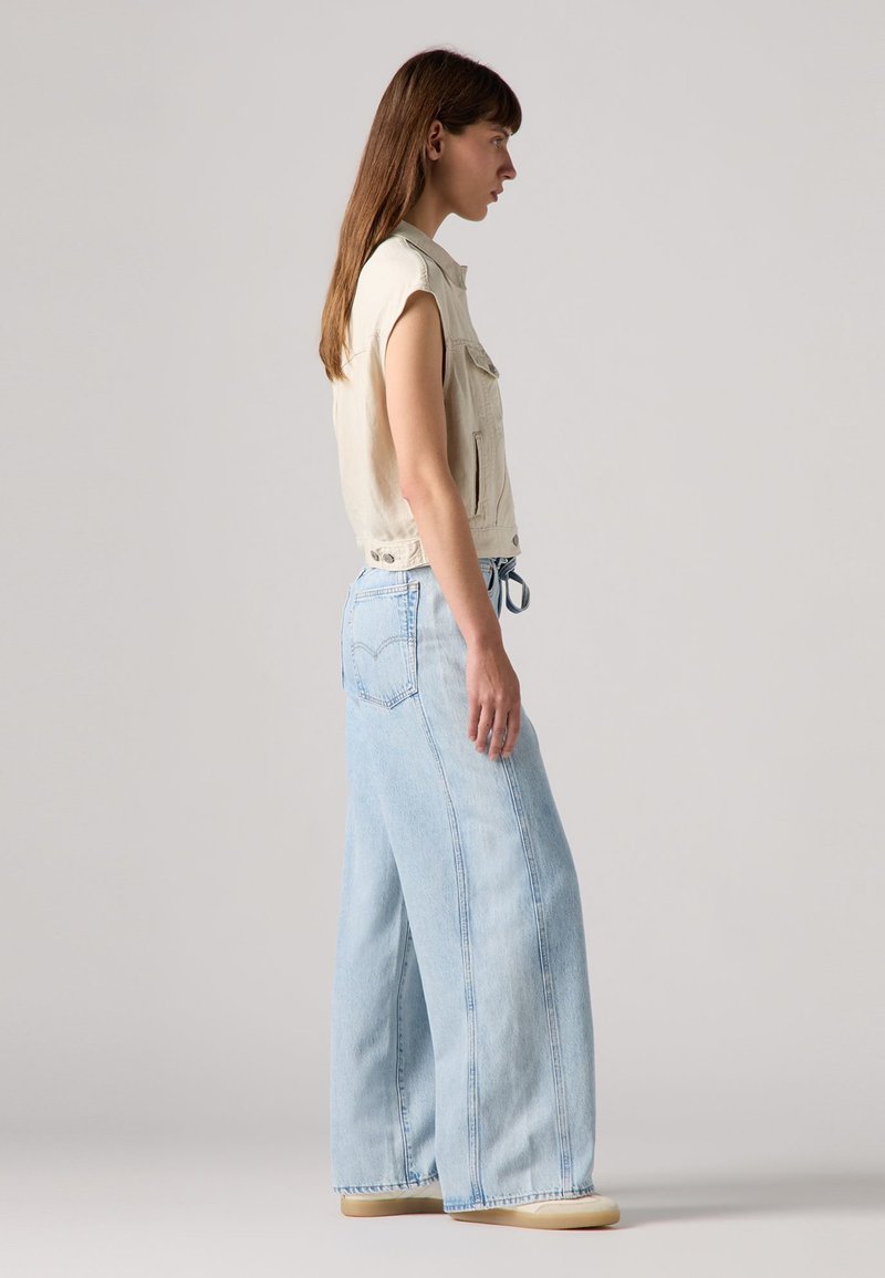 Femme se tenant de côté, portant un gilet sans manches beige et un jean large bleu clair avec des chaussures beiges sur un fond neutre.