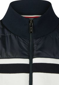 Marineblauwe kraagjas met een gevoerde textuur, voorzien van een ritssluiting aan de voorkant en horizontale witte en marineblauwe strepen over de borst.