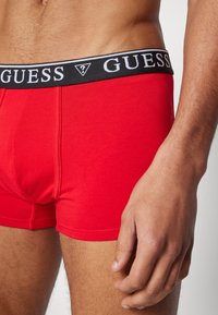 Crvene pamučne bokserice s crnim elastičnim pojasom na kojem je logotip "GUESS". Uvećani prikaz pokazuje glatku teksturu i oblik koji prianja.