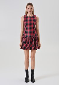 Donna che indossa un vestito senza maniche rosso e nero a quadri con orlo arricciato e stivaletti neri alla caviglia, in piedi davanti a uno sfondo chiaro e uniforme.