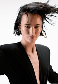 Prenda negra con escote en V profundo y mangas largas. La modelo tiene el cabello negro liso y lleva grandes pendientes de aro dorados. Fondo blanco liso.
