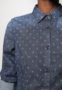 Mörkblå denimskjorta med ljusblåa prickar. Har en knapp-down krage och knappar fram, samt en slät, texturerad yta.