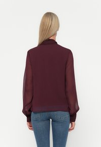 Camicia bordeaux con maniche lunghe trasparenti, colletto alto e polsini elasticizzati, abbinata a jeans blu di denim, vista da dietro.