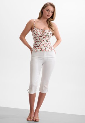 Femme debout, portant un pantalon blanc court, un haut sans manches à motifs cerises et des sandales dorées à lanières, sur un fond clair uni.