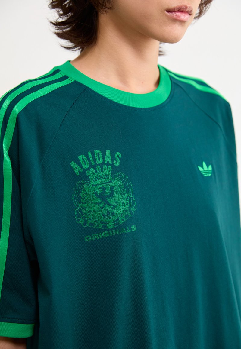 Teal groene oversized t-shirt met contrasterende groene strepen op de schouders, ronde halslijn en groene leeuw grafiek met de tekst "ADIDAS ORIGINALS".