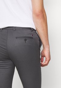 Šedé slim-fit kalhoty s kapsou na knoflík na zadní straně a malou logovou cedulkou. Tkanina vypadá hladce a je lehká. Pas má poutka na opasek.