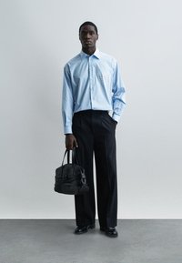 Άνδρας που στέκεται μπροστά σε απλό τοίχο, φορώντας ανοιχτό μπλε πουκάμισο με κουμπιά και μαύρο παντελόνι, κρατώντας μαύρη δερμάτινη τσάντα στο αριστερό χέρι.