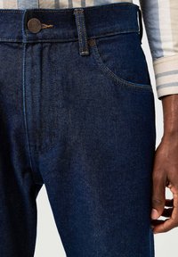 Jeans de mezclilla azul oscuro con corte recto, que presentan detalles de costura, un botón de metal en la cintura y un bolsillo frontal.