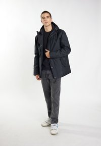 Giacca nera impermeabile con cappuccio, dotata di zip e bottoni a scatto. Indossata sopra una maglietta nera e abbinata a jeans grigi e sneaker bianche.
