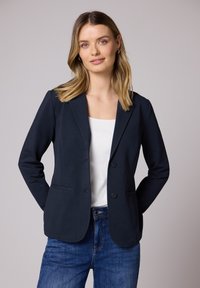 Jeune femme aux cheveux blonds portant un blazer bleu marine sur une chemise blanche et un jean bleu, debout devant un fond gris clair uni.