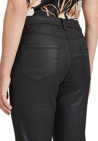 Schwarze Jeans mit einer glatten, glänzenden Oberfläche, ausgestattet mit zwei aufgesetzten Taschen auf der Rückseite und dezenten Nähdetails. Die Passform ist schmal am Bein.