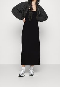 Svart ärmlös maxiklänning i kombination med en mörk bomberjacka och silverfärgade sportskor med gröna accenter. Slätt tyg, avslappnad look.