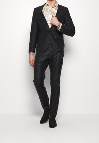 Abito nero texturizzato con una camicia oro a fantasia sotto, vestibilità su misura e scarpe nere. Dettagli notevoli includono una lieve lucentezza e rever affilati.