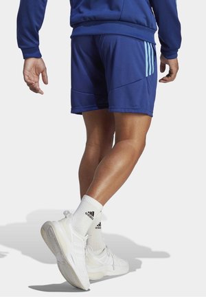 Man die blauwe sportshorts, een blauwe jas, witte Adidas-sokken en witte sneakers draagt, lopend met één voet omhoog.