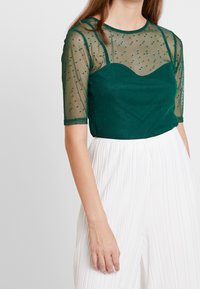 Top verde transparente con mangas cortas, patrón de puntos y escote corazón, superpuesto a una capa inferior verde sólida. Combinado con pantalones blancos con pliegues.