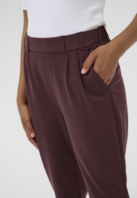 Pantaloni bordeaux con una vestibilità sartoriale, dotati di pieghe, tasche laterali e una texture del tessuto liscia. Mani appoggiate nelle tasche.