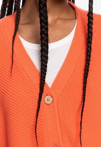 Strukturierte orangefarbene Strickjacke mit V-Ausschnitt und Holzknöpfen, getragen über einem weißen Hemd.