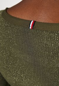 Olijfgroen geribbeld topje met een metallic glans, met een ronde halslijn en een klein accent in rood, wit en blauw aan de achterkant.