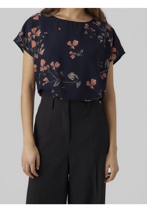 Vrouw draagt een marineblauwe blouse met roze bloemenprint en zwarte wijde broek met hoge taille tegen een effen achtergrond.