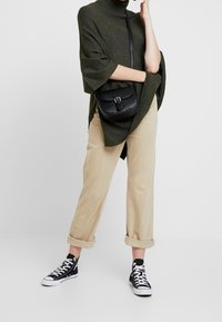 Oliwkowo-zielony, oversize'owy sweter z dzianiny z wysokim kołnierzem, zestawiony z beżowymi spodniami z podwiniętymi nogawkami i czarnymi wysokimi trampkami. Czarna torba na ramię.