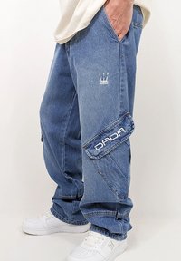 Blue denim cargo pants featuring elastic cuffs, side pockets, and embroidered "DADA" logo. Includes a small crown emblem on the thigh.