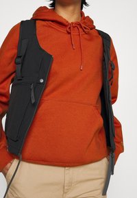 Chaleco negro con múltiples bolsillos y correas ajustables, llevado sobre una sudadera naranja con capucha. La textura parece suave con un acabado mate.