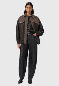 Vrouw in een oversized bruin en zwart jasje, met een luipaardprint top, zwarte leren broek en zwarte schoenen, staande met één hand in de zak.