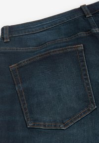 Jeans din denim de un albastru închis au o buzunar din spate cu cusături portocalii și o textură netedă, surprinzând luciul și structura subtilă a materialului.