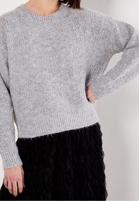 Femme portant un pull en tricot gris clair texturé à manches longues et une jupe plissée noire, debout avec une main sur la hanche.