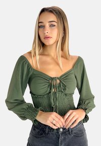 Olivgrön blus med knytframslutning, smockad byst och puffärmar. Modellen bär högmidjade svarta jeans som framhäver detaljernas i blusen.