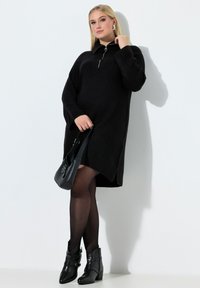 Zwarte geribbelde trui-jurk met een halve ritssluiting en zijsplitsingen. Gecombineerd met zwarte enkellaarsjes en een kleine zwarte handtas. Gestructureerde stof.