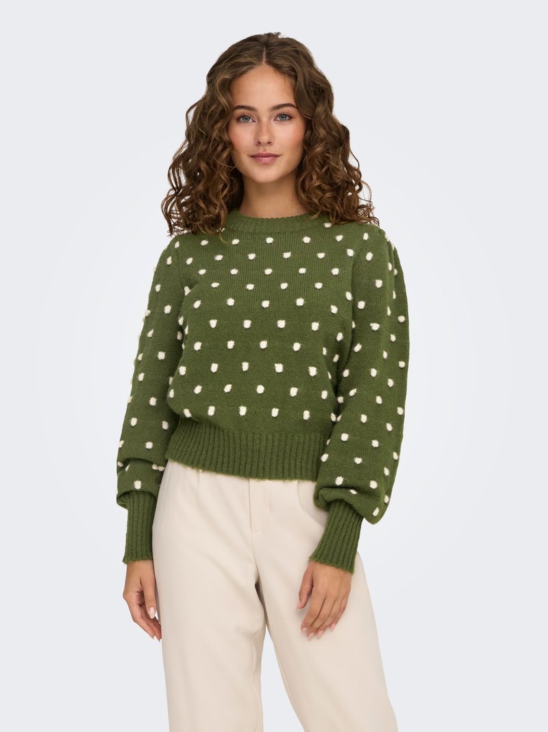 Femme aux cheveux bouclés portant un pull vert à pois blancs et un pantalon crème taille haute, debout devant un fond uni.