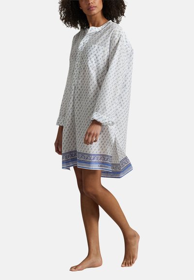 Femme debout pieds nus portant une robe blanche ample à manches longues avec de petits motifs floraux bleus et un ourlet décoratif bleu.