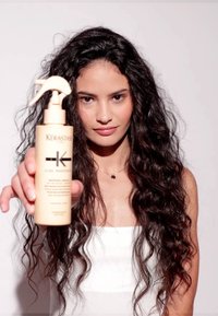 Producto capilar Kérastase en un envase beige con un pulverizador, que presenta texto y detalles de etiqueta en negro. El fondo es liso y blanco.