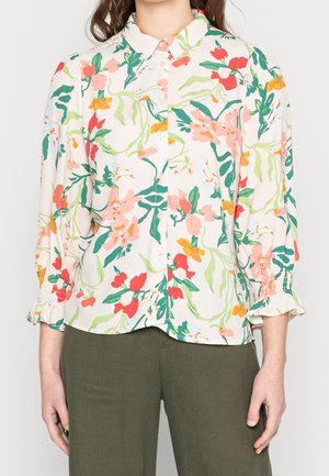 Kobieta ubrana w koszulę na guziki z różowymi, czerwonymi, pomarańczowymi i zielonymi kwiatowymi wzorami oraz oliwkowo zielone spodnie, stoi na jednolitym tle.